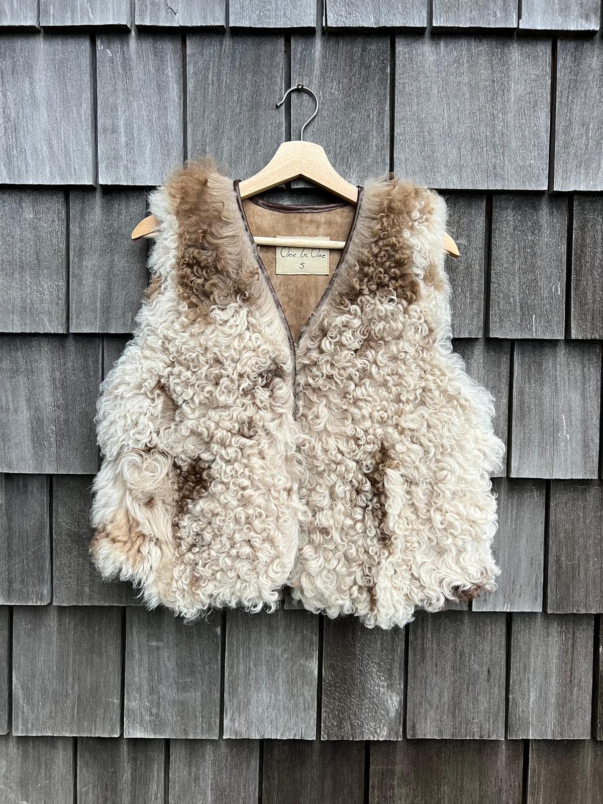 Cream curly sheepskin vest with caramel tips on a wooden hanger against grey shingles
